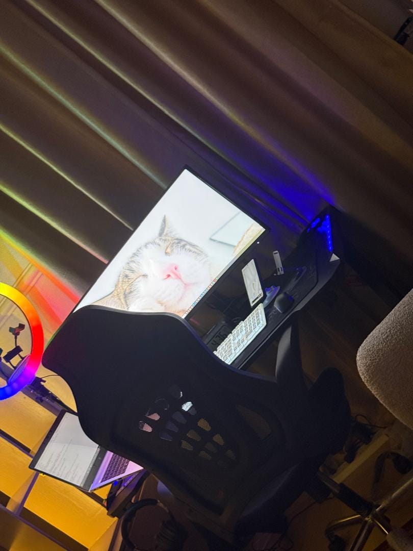 Angled view of the same workspace showing the monitor, keyboard, mouse, laptop and desk chair under purple and blue lighting