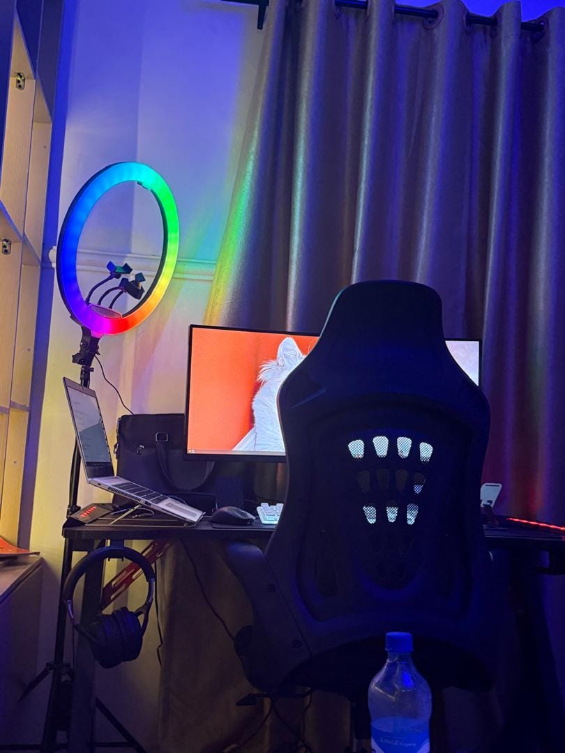 JOEWALL DEV workspace: a desk with a monitor, laptop on a stand, keyboard and headphones, lit by a rainbow ring light and blue accent lighting